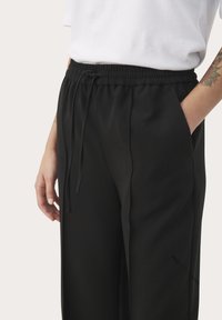 Černé kalhoty s elastickým pasem, detaily na šňůrku a bočními kapsami, vyrobené z měkké látky s hladkým povrchem. Jednoduchý design.