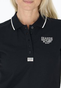 Marine polo med hvid kravekant, fem-knaps placket, og broderet "Seaside Nautical Legacy"-logo. Har et vævet mærke.