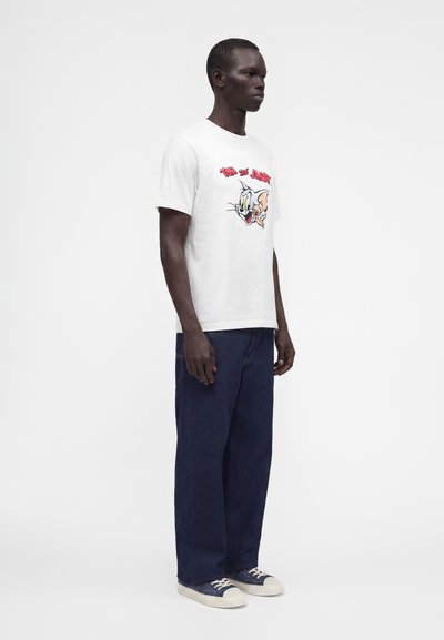 Un homme porte un t-shirt blanc Tom et Jerry, un jean bleu foncé et des baskets en toile bleu marine, debout sur un fond blanc uni.