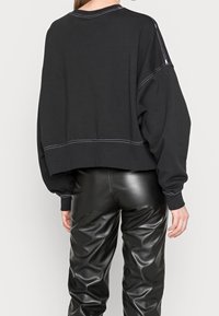 Sudadera negra corta con mangas extragrandes, costuras blancas en contraste, combinada con pantalones de cuero negro brillante. Textura de tela suave visible.