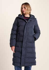 Woman wearing a long, navy blue puffer coat with hood, standing with one hand in pocket, looking slightly to the side against a plain background.