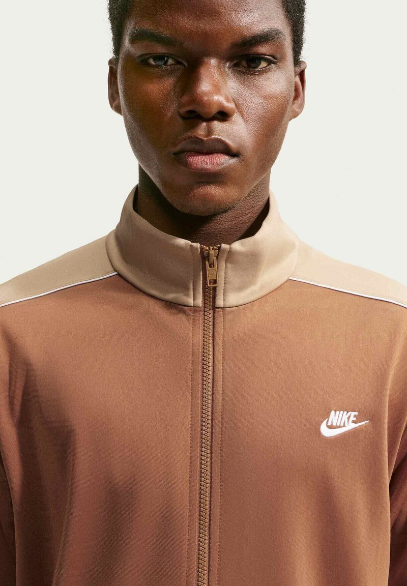Ung mand iført en brun og beige Nike jakke med lynlås og hvidt logo på venstre bryst, som vender fremad med et neutralt udtryk.