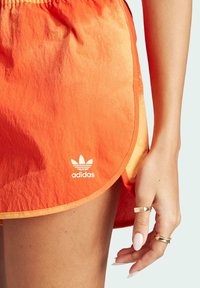 Atletska krila v oranžni gradient barvi, izdelana iz lahkega materiala. Ima ukrivljen rob in belo Adidas logo na spodnjem levem delu.