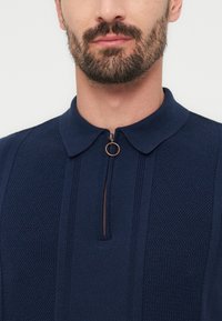 Άνδρας με περιποιημένο γένι φοράει μπλε ναυτικό πλεκτό πόλο με υφή, εμπρόσθιο φερμουάρ και στρογγυλό μεταλλικό τράβηγμα.