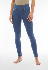 Mujer vistiendo jeggings azules de talle alto con bolsillos delanteros, de pie descalza contra un fondo blanco.