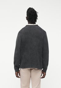 Tamsiai pilkas megztinis iš mezgimo su ripsuota tekstūra, apvalia apykakle ir kontrastuojančia šviesia apdaila prie kaklo. Laisvo silueto su ilgomis rankovėmis.