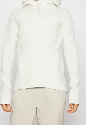 Sudadera blanca con cremallera y capucha ajustable, textura suave, mangas largas, bolsillos frontales y puños ajustados. Combinada con pantalones beiges.