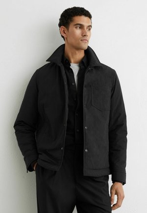 Schwarze, strukturierte Jacke mit hohem Kragen, Knopfverschluss und einer Seitentasche; getragen über einem schwarzen Hemd mit grauen Akzenten.