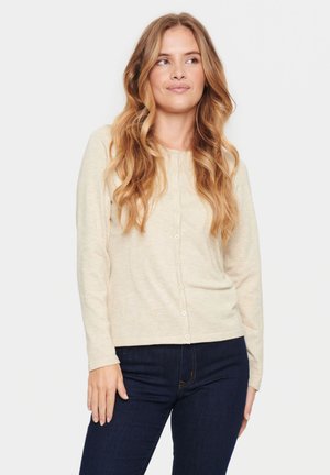 Beige Langarm-Cardigan mit Rundhalsausschnitt, vorderem Knopfverschluss und weicher Textur, kombiniert mit dunkelblauen Jeans.