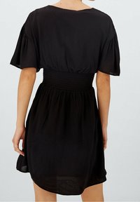 Nainen, jolla on päällään musta mekko, jossa on smockattu vyötärö ja väljät lyhyet hihat, näkyy takaa yksinkertaista taustaa vasten.