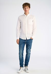 Witte longsleeve shirt met een knoopsluiting, enkele borstzak, gecombineerd met blauwe slim-fit jeans en witte sneakers met kleurrijke veters.