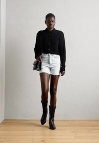 Camicia nera con bottoni, pantaloni corti in denim bianco strappati e stivali neri fino al ginocchio. La modella porta una piccola borsa a tracolla, su un pavimento in legno.