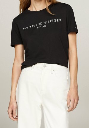 Kobieta nosząca czarny T-shirt Tommy Hilfiger z logo i tekstem "Est. 1985", zestawiony z białymi spodniami z wysokim stanem na jednolitym tle.