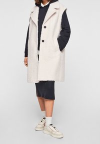 Cappotto senza maniche di un beige chiaro realizzato in un tessuto texturizzato e morbido, con due tasche frontali e bottoni neri. Indossato sopra un hoodie blu navy e una gonna.