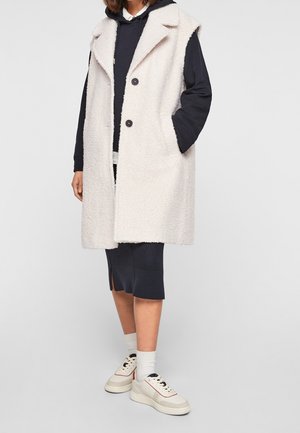 Manteau sans manches beige clair en tissu texturé et duveteux, avec deux poches avant et des boutons noirs. Porté par-dessus un hoodie bleu marine et une jupe.