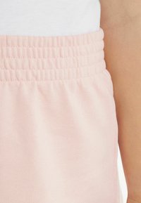 I pantaloni della tuta rosa chiaro presentano un cintura elasticizzata arricciata e un tessuto morbido e liscio, offrendo una vestibilità comoda e una texture piacevole.