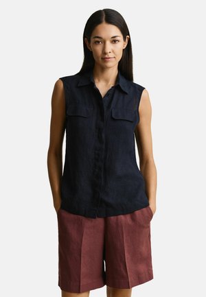 Frau mit langem, dunklem Haar, die ein ärmelloses, schwarzes Hemd mit Kragen und lockere, knieumspielende Shorts in Bordauxrot trägt, steht mit den Händen in den Taschen.