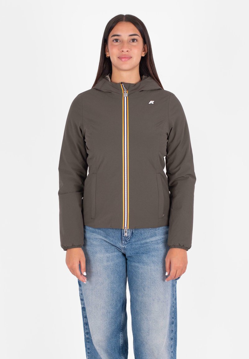 Jonge vrouw in een donkergrijze hoodie met rits en lichtblauwe spijkerbroek, staand tegen een effen witte achtergrond.