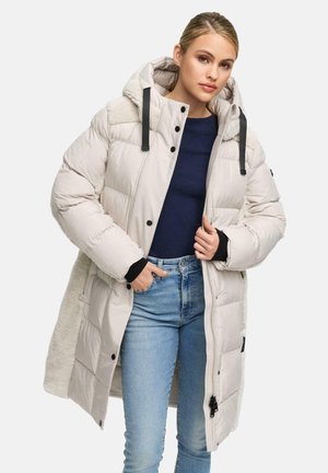 Lichtbeige pufferjas met capuchon, met zwarte accenten, een ritssluiting aan de voorkant en een bontvoering. Ggedragen over een donkere top en jeans.