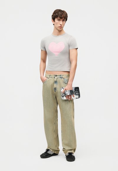Νέος άνδρας που φοράει γκρι εφαρμοστό crop top με ροζ καρδιά, μπεζ παντελόνι με φαρδιές παντελόνες, μαύρα αθλητικά παπούτσια και κρατάει μεταλλική τσάντα clutch.