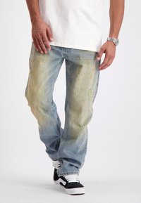 Man in versleten, distressed blauwe jeans, zwart-witte sneakers, wit T-shirt en zilveren polshorloge die tegen een witte achtergrond loopt.
