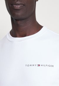 T-shirt en coton blanc avec un col rond. Présente le logo "TOMMY HILFIGER" en texte noir, rouge et bleu sur la partie gauche de la poitrine. Texture lisse.
