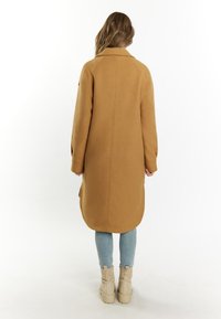 Cappotto lungo marrone con colletto, texture liscia e tasche laterali. Abbinato a jeans azzurri e stivaletti beige. Vista posteriore mostrata.