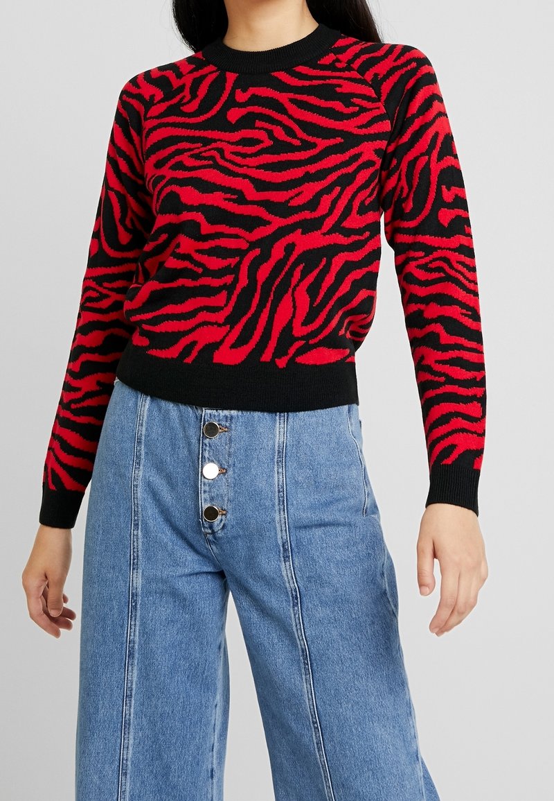 Červeno-černý svetr s pruhy ve vzoru zebry, kulatým výstřihem a žebrovanými manžetami. Spárován s vysokými světle modrými širokými džíny s knoflíky.