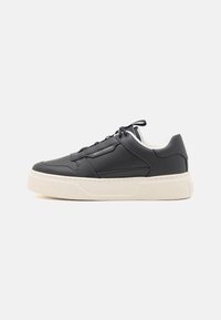 Sorte lærersneakers med en teksturert overflate, hvit gummiklarg, og flate lisser. Har subtil merkevaremerking på siden. Lave topper.