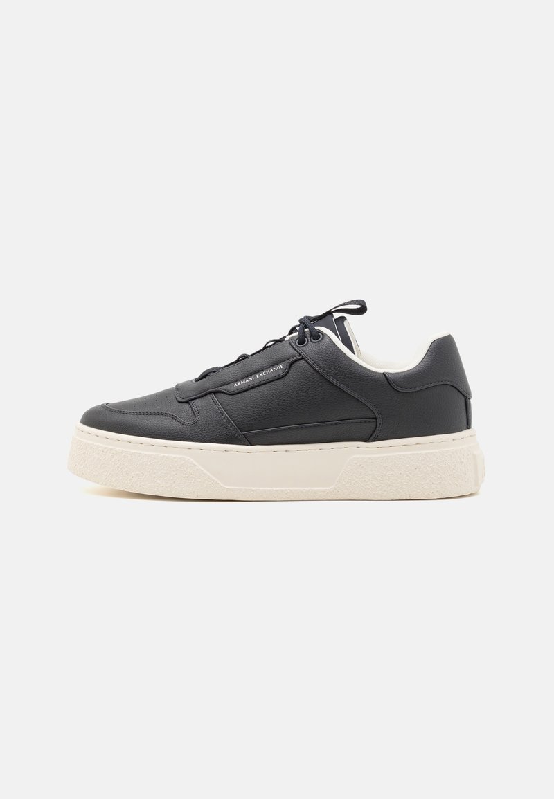 Sorte lærersneakers med en teksturert overflate, hvit gummiklarg, og flate lisser. Har subtil merkevaremerking på siden. Lave topper.