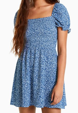 Woman wearing a blue and white floral short dress with puffed short sleeves and a square neckline.