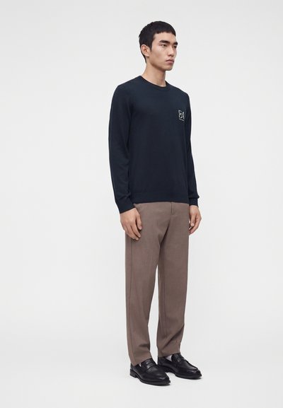 Homme debout portant une chemise noire à manches longues avec un petit logo, un pantalon marron et des chaussures en cuir noires sur fond blanc uni.