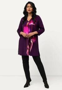 Tunic viola con stampa floreale in rosa e bianco, scollatura a V, maniche a tre quarti, spacchi laterali, abbinata a pantaloni neri e tacchi.