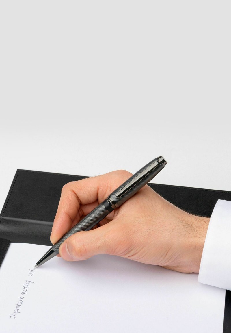 Un stylo noir tenu dans une main, écrivant sur du papier blanc. L'arrière-plan comprend un dossier noir, avec une texture visible et une finition lisse.
