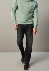 Sudadera verde claro con un bolsillo en el pecho con cremallera, combinada con jeans oscuros y zapatillas negras. Tela suave y diseño ajustado.