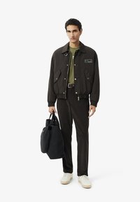 Giovane uomo che indossa un giaccone marrone scuro e pantaloni abbinati, con una maglia verde, scarpe da ginnastica bianche, mentre porta una grande borsa a tracolla nera, in piedi contro uno sfondo bianco.