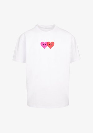 Baltas kokvilnas T-krekls ar centrālu grafiku, kurā attēloti divi smaidīgi sirdis – viena rozā un otra sarkana, ar izteiksmīgām acīm.