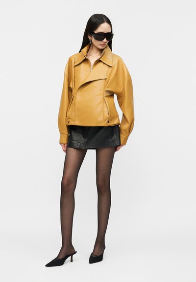 Woman wearing oversized mustard leather jacket, black leather mini skirt, sheer black tights, black pointed heels, and large black sunglasses.