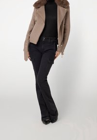 Veste en laine marron avec col en fourrure, pantalon cargo noir ajusté et escarpins noirs pointus. Fermetures éclair et boutons visibles sur la veste.