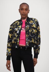 Black bomber jacket featuring gold and white ornate patterns, with ribbed cuffs and waistband. Paired with a pink fitted sweater.
