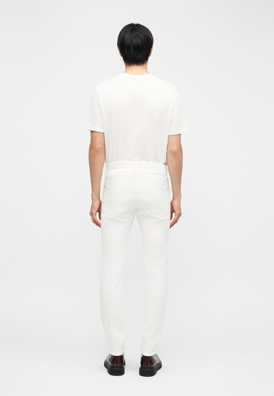 Homme debout, dos tourné, portant une chemise blanche à manches courtes, un pantalon blanc et des chaussures marron foncé sur un fond blanc uni.