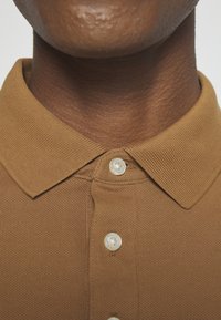 Polo shirt marron en tissu texturé et côtelé, avec un col pointu et trois boutons blancs au niveau du col.