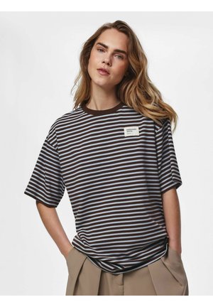 Jeune personne aux longs cheveux portant une chemise rayée marron et bleu clair avec un patch "ESSENTIALS" et un pantalon beige, les mains dans les poches.
