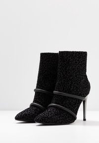 Stivaletti neri in suede con texture leopardata, punta affilata e sottili cinturini in metallo nero attorno alla parte centrale, dotati di alti tacchi a stiletto.