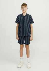 Chemise à manches courtes à boutons et shorts assortis en bleu marine avec des rayures verticales blanches, fabriqués en tissu léger. Baskets blanches aux pieds.