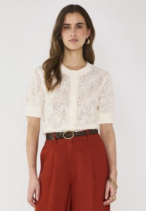 Giovane donna con cardigan bianco a maniche corte a bottoni con motivo, pantaloni a vita alta color ruggine, cintura intrecciata e orecchini a cerchio dorati.