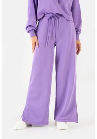 Fioletowe spodnie o szerokich nogawkach wykonane z miękkiego materiału, z elastycznym pasem i sznurkiem. Posiadają boczne rozcięcia na dole i są zestawione z białymi sneakersami.