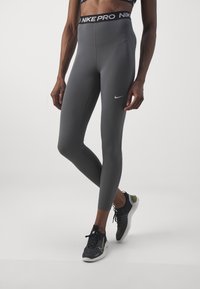 Šedé legíny s vysokým pasem a jednoduchým designem. Mají černý pás "NIKE PRO" a bílé logo Nike na dolní části nohy.