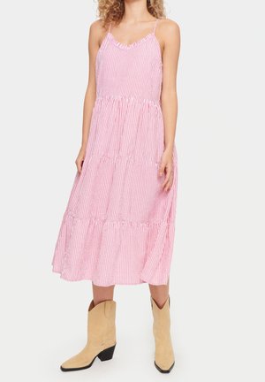 Vrouw draagt een mouwloze midi-jurk met roze en witte strepen en lagen, gecombineerd met cognackleurige suède cowboylaarzen op een effen achtergrond.