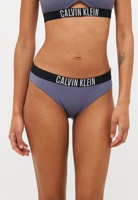 Vijolične bikini hlačke z črnim elastičnim pasom, ki ima belo blagovno znamko "CALVIN KLEIN". Zasnovane za tesno prileganje.